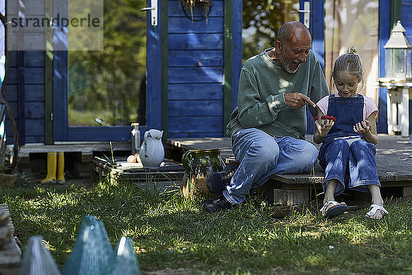 Großvater und Enkelin mit Himbeeren auf ihren Fingerspitzen auf der Terrasse im Garten sitzend