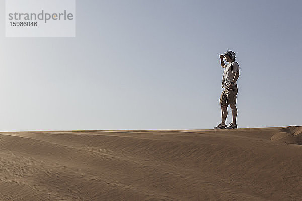 Männlicher Tourist  der seine Augen abschirmt  während er auf einer Sanddüne gegen den klaren Himmel in Dubai  Vereinigte Arabische Emirate  steht