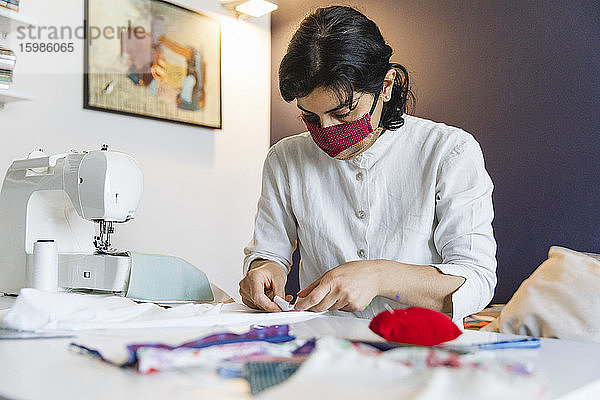 Selbstbewusste Frau mit Gesichtsmaske  während sie zu Hause an der Nähmaschine sitzt