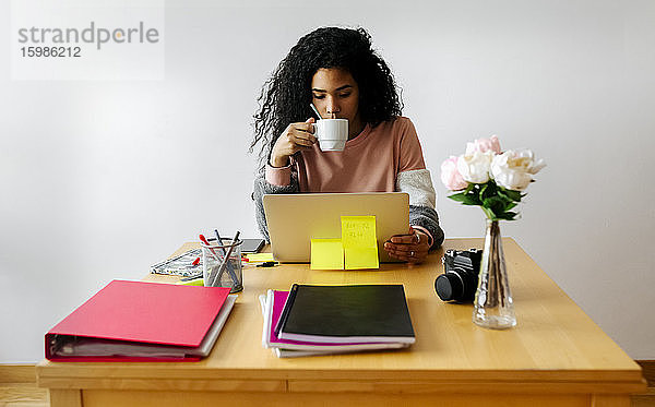 Junge Frau arbeitet von zu Hause aus mit Laptop und trinkt Kaffee