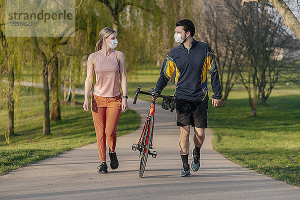 Junges Paar  das während der Coronavirus-Pandemie mit dem Fahrrad auf dem Fußweg im Park spazieren geht