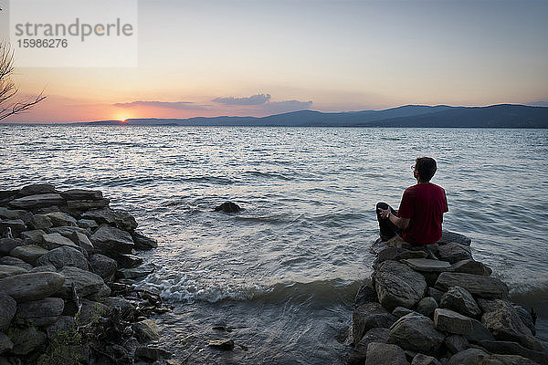 Rückansicht eines reifen Mannes  der bei Sonnenuntergang auf einem Felsen sitzend den Trasimenischen See betrachtet  Isola Maggiore  Italien