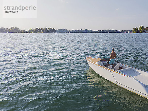 Deutschland  Bayern  Mann ohne Hemd steht auf einem Motorboot im Chiemsee und bewundert die Landschaft in der Ferne
