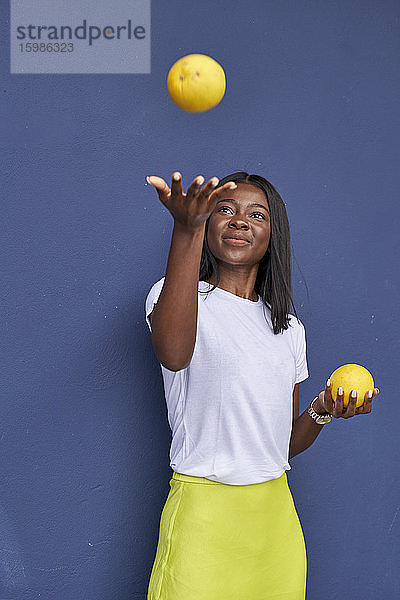 Porträt einer glücklichen jungen Frau  die mit zwei Orangen vor einem blauen Hintergrund jongliert