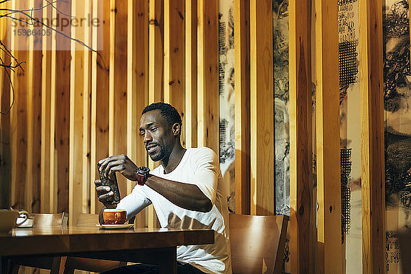Lächelnder junger Mann  der sein Smartphone benutzt  während er mit einem Kaffee auf einem Tisch an der Wand eines Cafés sitzt