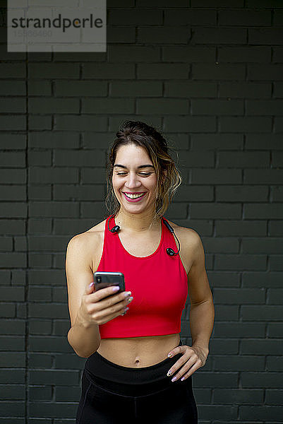 Lächelnde  selbstbewusste Sportlerin  die ein Smartphone benutzt  während sie an einer Mauer steht