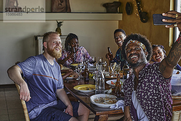Lächelnder Mann  der ein Selfie mit Freunden macht  die während des Mittagessens am Esstisch sitzen