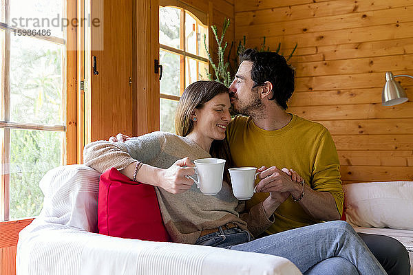 Romantischer Freund hält Kaffeetasse und küsst Frau auf die Stirn  während er in einer Blockhütte sitzt