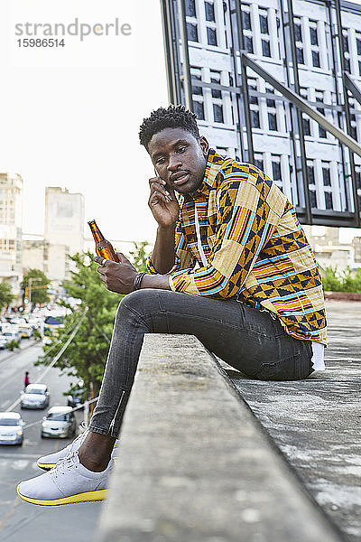 Junger Mann in gemustertem Hemd sitzt auf einer Dachterrasse in der Stadt und telefoniert  Maputo  Mosambik