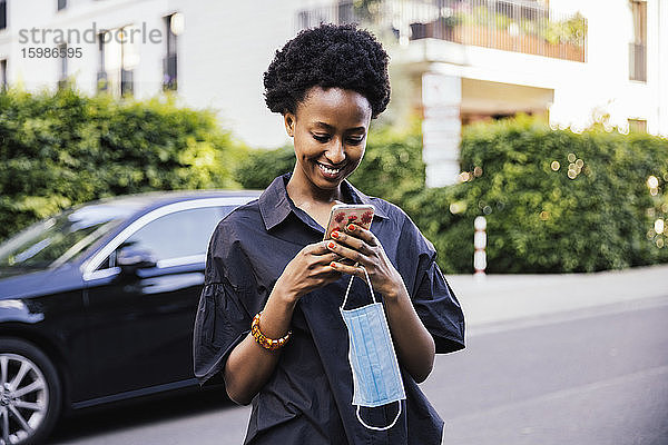 Porträt einer lächelnden jungen Frau mit Schutzmaske  die auf der Straße steht und auf ihr Mobiltelefon schaut