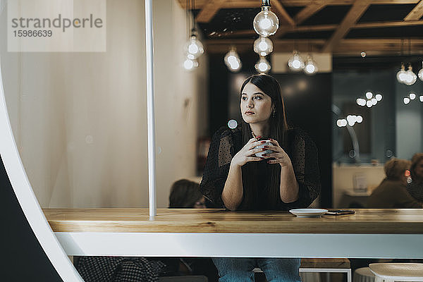 Schöne junge Frau schaut weg  während sie mit einer Kaffeetasse im Café sitzt und nachdenkt