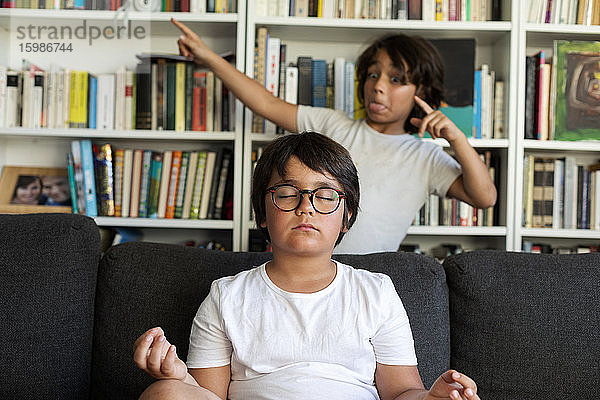 Junge sitzt auf der Couch und meditiert  sein Bruder ärgert ihn