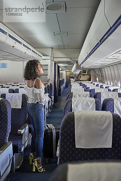 Junger weiblicher Passagier auf der Suche nach einem Sitzplatz in voller Länge  während er im Gang eines Flugzeugs steht