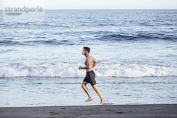 Shirtless Mitte erwachsenen Mann trägt Gesichtsmaske beim Laufen am Strand bei Sonnenuntergang