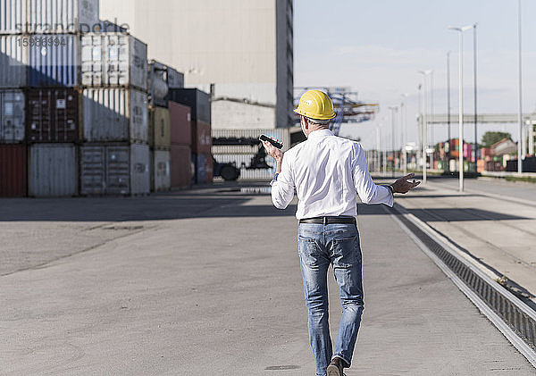 Rückenansicht eines Geschäftsmannes mit Schutzhelm  der auf einem Industriegelände ein Mobiltelefon benutzt