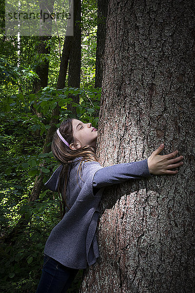 Nettes Grundschulmädchen umarmt Baumstamm im Wald