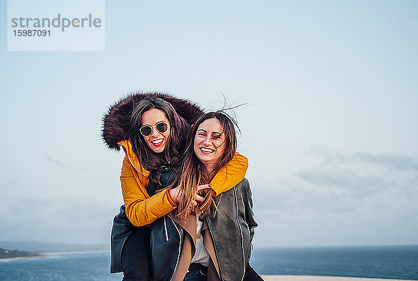 Frau  die eine fröhliche Freundin huckepack nimmt  während sie sich am Strand gegen den Himmel in Tarifa  Spanien  vergnügt