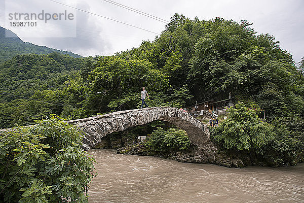 Georgien  Adscharien  Makhuntseti  Junger Mann überquert Steinbogenbrücke über braunen Fluss