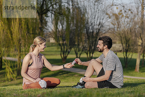 Ein lächelnder Mann gibt einer Frau Handdesinfektionsmittel  während er im Park auf dem Rasen sitzt
