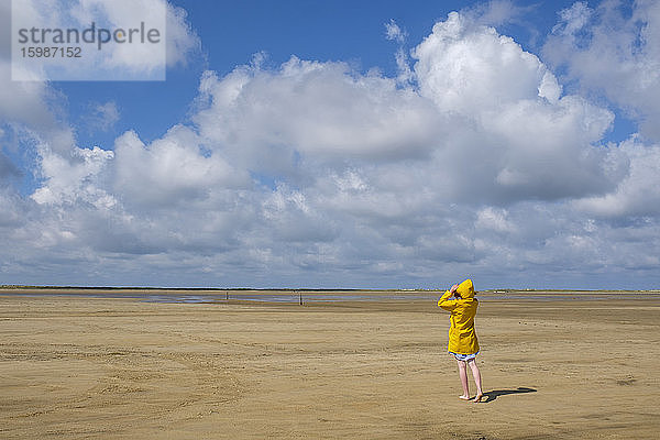 Volle Länge Rückansicht von Teenager-Mädchen trägt gelben Regenmantel beim Gehen auf Sand am Strand während des sonnigen Tages