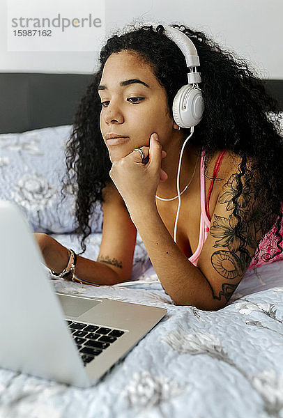 Schöne junge Frau liegt zu Hause im Bett  trägt Kopfhörer und benutzt einen Laptop