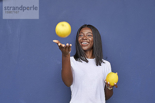 Porträt einer glücklichen jungen Frau  die mit zwei Orangen vor einem blauen Hintergrund jongliert