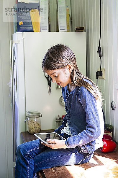 Mädchen sitzt auf der Arbeitsplatte in der Küche und schaut auf ein digitales Tablet