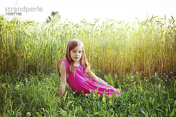 Porträt eines jungen Mädchens in einem leuchtend rosa Kleid  das vor einem Roggenfeld im Sommer sitzt