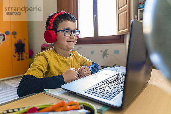 Lächelnder männlicher Schüler  der während des Hausunterrichts über Kopfhörer zuhört und auf seinen Laptop schaut