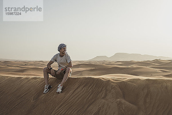 Männlicher Tourist schaut weg  während er auf Sanddünen in der Wüste von Dubai  Vereinigte Arabische Emirate  sitzt