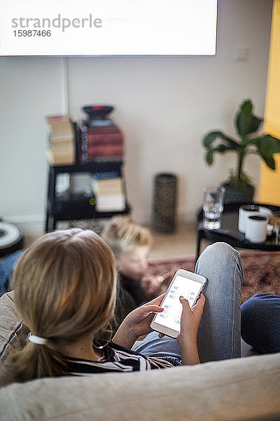 Rückansicht eines Mädchens  das ein Smartphone benutzt  während es zu Hause auf dem Sofa sitzt