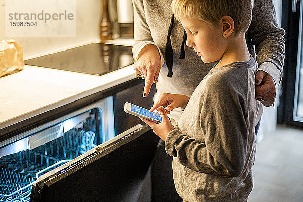 Junge lernt von der Mutter  während er eine mobile App über den Geschirrspüler in der Küche im Smart Home benutzt