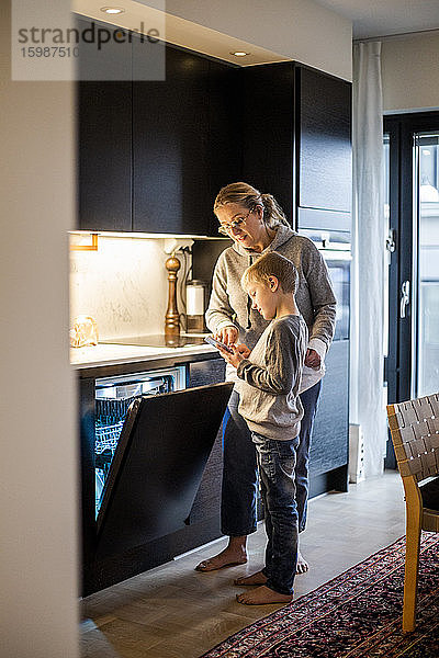 Mutter bringt Sohn bei  eine mobile App zu benutzen  während er zu Hause in der Küche den Geschirrspüler bedient