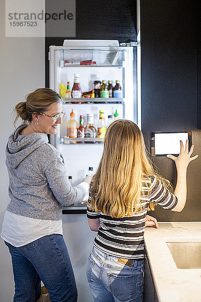 Tochter benutzt digitales Tablett  während die Mutter im Smart Home am Kühlschrank steht