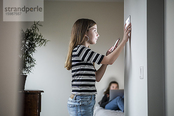 Mädchen hält Mobiltelefon in der Hand  während sie ein digitales Tablet an der Wand benutzt  während die Schwester im Hintergrund im Smart Home sitzt