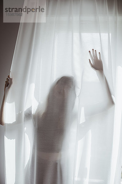 Junge Frau versteckt sich hinter weißem Vorhang