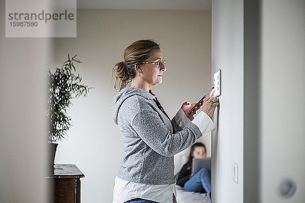 Mutter hält Smartphone in der Hand  während sie ein digitales Tablet an der Wand hält  während die Tochter im Hintergrund im Smart Home ist