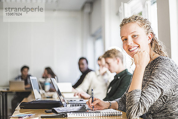 Porträt einer lächelnden jungen Frau am Schreibtisch im Klassenzimmer