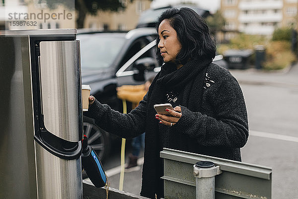Mittlere erwachsene Frau mit Smartphone an der Ladestation für Elektroautos