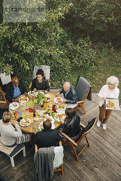 Schrägaufnahme einer Frau  die älteren Freunden während einer Gartenparty im Hinterhof am Esstisch sitzt