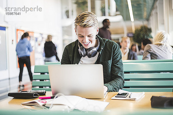 Lächelnder junger männlicher Student benutzt Laptop in der Cafeteria der Universität
