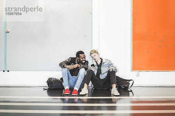 Junger Mann zeigt einem Freund sein Handy  während er im Flur der Universität sitzt
