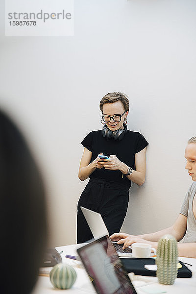 Lächelnder junger geschlechtsgemischter Geschäftsmann benutzt Smartphone  während er sich im Sitzungssaal an die Wand lehnt