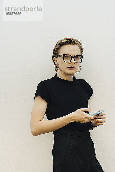 Selbstbewusster androgyner Unternehmer steht mit Smartphone an der Wand im Sitzungssaal