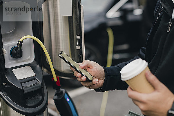 Mittelsektion eines Mannes  der ein Smartphone an einer Ladestation für Elektroautos benutzt