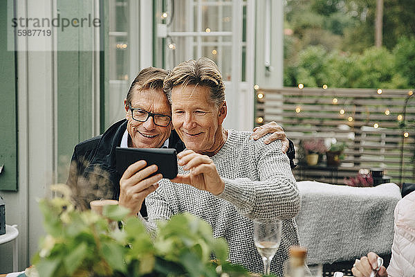 Lächelnder Mann zeigt Smartphone mit Freund  während er während der Party im Garten sitzt