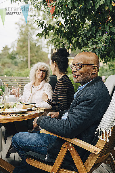 Lächelnder kahlköpfiger älterer Mann sitzt auf Stuhl und genießt Dinnerparty von Freunden im Hinterhof
