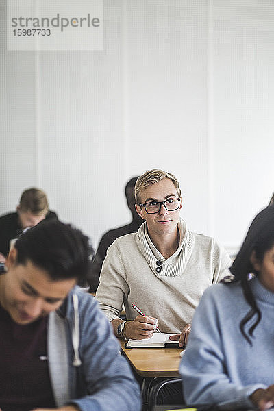 Junger Mann schreibt in Buch am Schreibtisch im Klassenzimmer