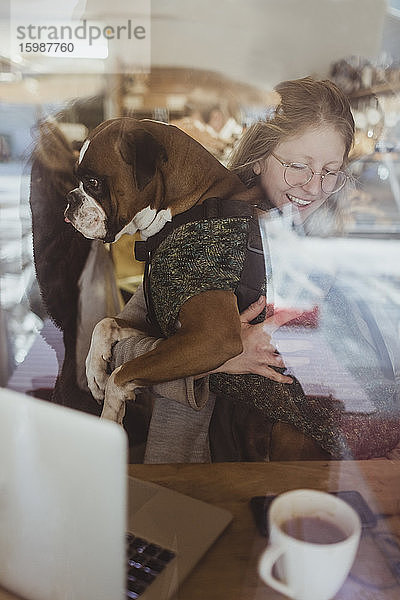 Lächelnde junge Frau sitzt mit Boxerhund durch Café-Fenster gesehen