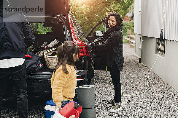 Lächelnde Frau lädt Auto auf  während die Familie Gepäck in den Kofferraum lädt